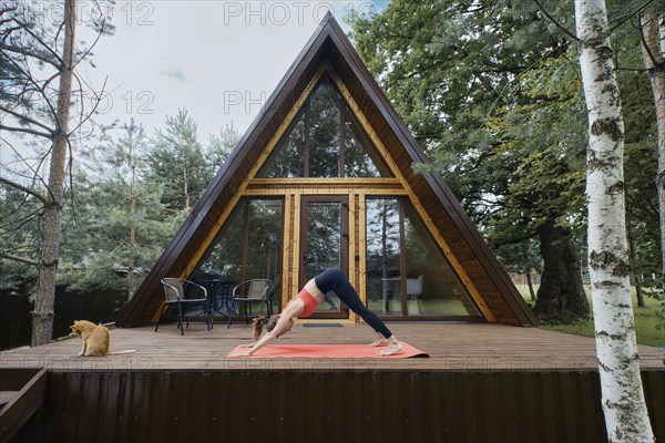 Flexible woman standing in dog's face-down pose on wooden terrace of her summer cabin
