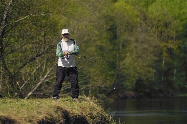 Fisherman throwing the bait into river standing on river bank in sunny day with green forest in
