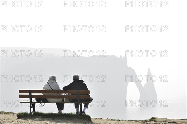 Elderly tourists sitting on bench looking at the Porte D'Aval, a natural arch in the chalk cliffs