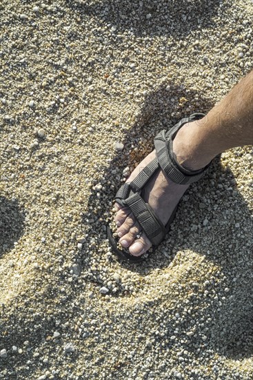 Close-up of man's foot in sandal standing on beach with coarse sea sand grains