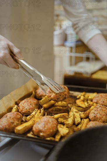 Closeup view of lifting meat patty from baking tray with potato wedges with kitchen tongs