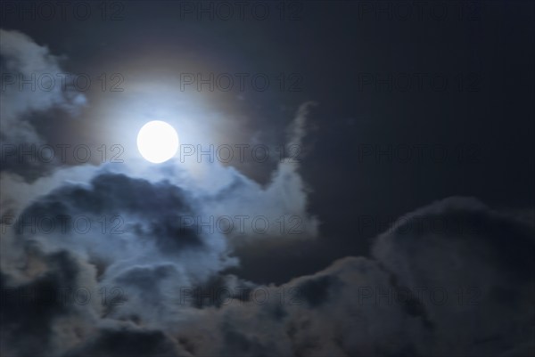 The full moon shines brightly in a cloudy, stormy, black sky and has a halo in rainbow colours