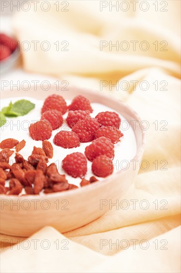 Yogurt with raspberry and goji berries in ceramic bowl on white concrete background and orange