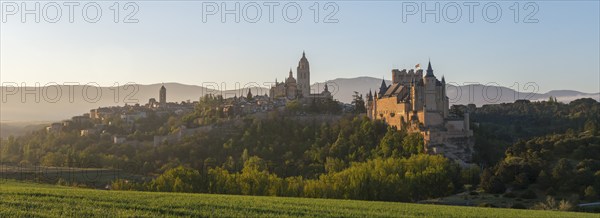 Panoramic view of a historic castle and town surrounded by green fields and mountains at sunrise,