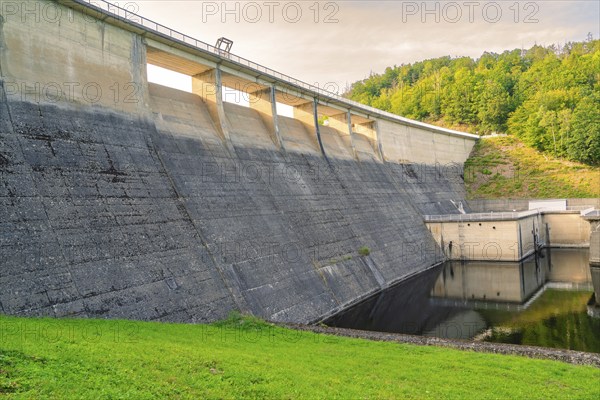 An imposing concrete dam with flowing water, embedded in a green hilly landscape,