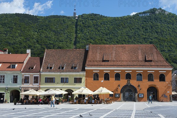 A lively town square in front of historic buildings with cafés at the foot of a green mountain,