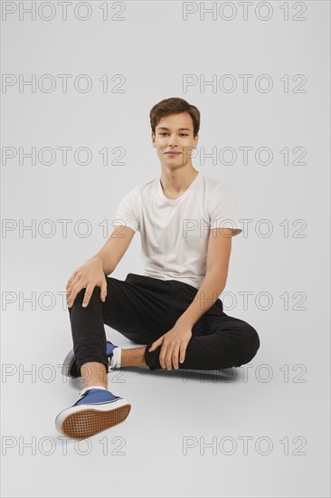 Cheerful young handsome man in black trousers and white shirt sits on the floor in studio