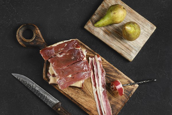 Cured duck meat on a wooden board next to two ripe pears. A knife rests nearby, highlighting the
