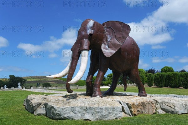 Metal sculpture of an elephant on a rock foundation in a green park under a blue sky with white