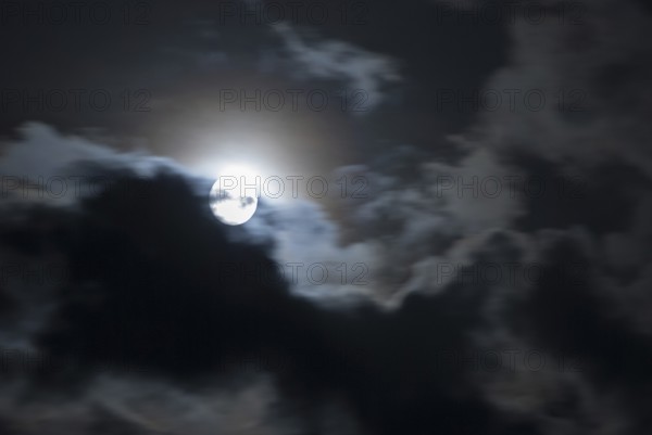 The full moon shines brightly in a cloudy, stormy, black sky and has a halo (corona),