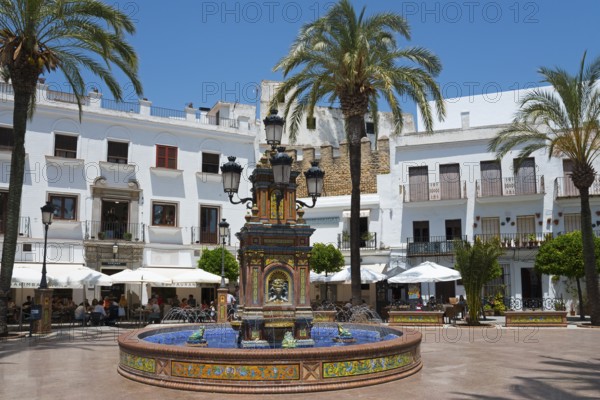 A central fountain in a sunny plaza surrounded by white buildings and palm trees, fountain with