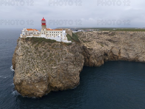 A red lighthouse and white buildings stand on an imposing cliff with a wide view of the ocean,