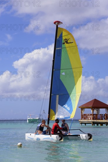 People on a colourful sailing boat in the clear sea near a jetty with a pavilion under a blue sky,