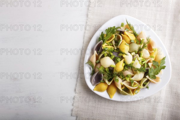 Conchiglie colored pasta with fresh greengrocery on a linen tablecloth on white wooden background.