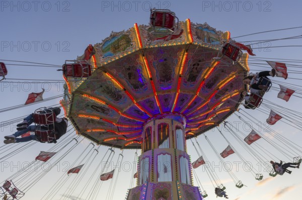 A colourfully illuminated carousel with swinging seats against an evening sky, wave flight, chain