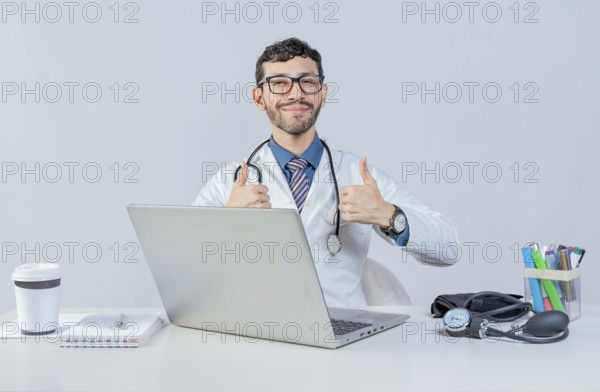 Smiling doctor sitting with laptop giving thumbs up. Male doctor with laptop showing thumbs up in