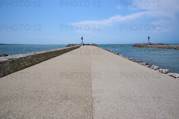 A long concrete path leads to the sea, flanked by breakwaters under a sunny, cloudy sky, summer, Le