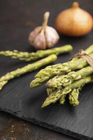 Bunch of fresh green asparagus, garlic, onion on slate board on black concrete background. Side