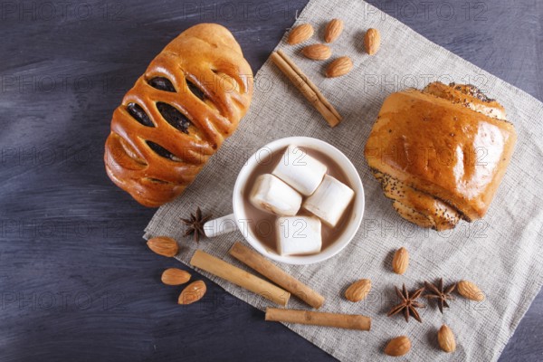 A cup of hot chocolate with marshmallow, buns and spices on linen napkin and black wooden