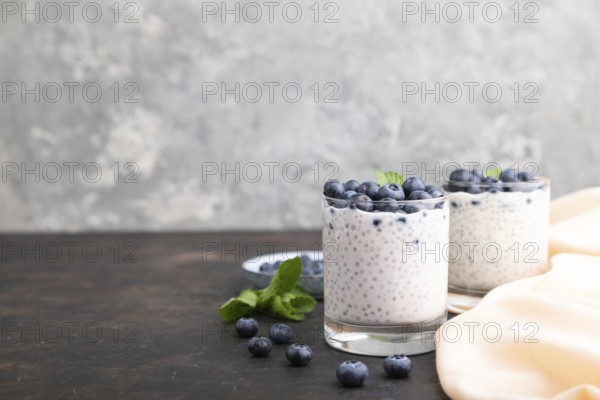 Yogurt with blueberry and chia in glass on black concrete background and orange linen textile. Side