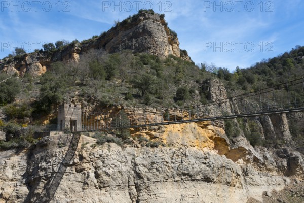 A rocky mountain landscape with dense vegetation under a bright blue sky, suspension bridge,