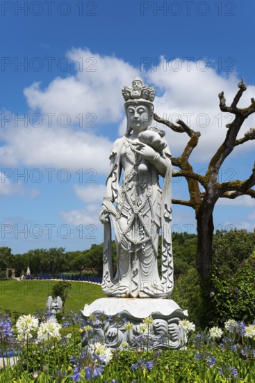 A stone Buddha statue surrounded by trees and flowers under a blue sky with clouds, Bacalhôa,