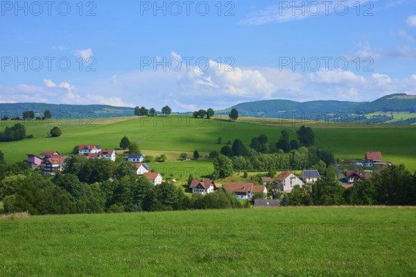 Small village nestled in a hilly landscape with green meadows and trees under a blue sky,