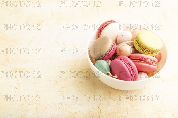 Multicolored macaroons and chocolate eggs in ceramic bowl on beige concrete background. side view,