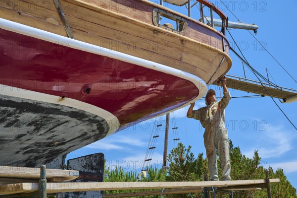 A worker works on the red hull of a wooden sailing ship in the dry dock, Patmos Marine, boatyard,