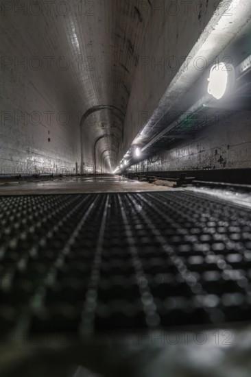 Long concrete tunnel with grid floor and damp surfaces, illuminated by lamps on the sides, Rappbode