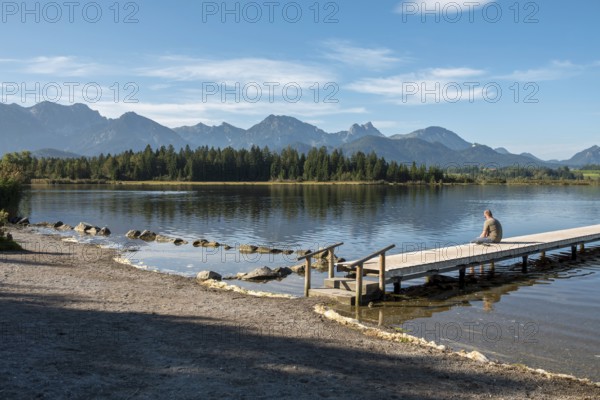 Person sitting on a wooden footbridge looking at a calm lake with mountains in the background,