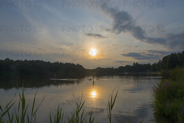 Calm lake at sunset, surrounded by trees and reeds, with a clear sky and scattered clouds, Oleš
