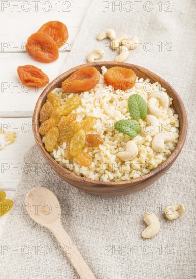 Bulgur porridge with dried apricots, raisins and cashew in wooden bowl on a white wooden background