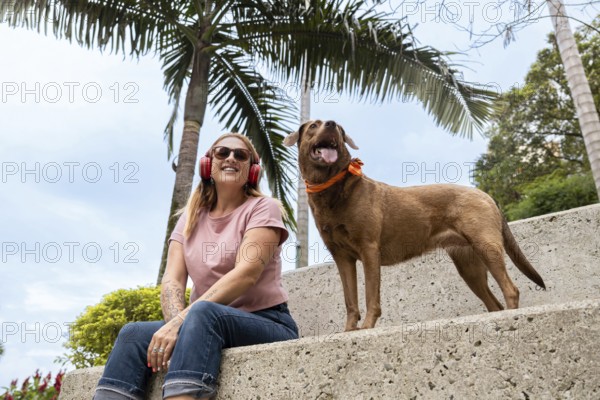 A smiling woman listens to music while sitting next to her alert dog wearing a colorful bandana
