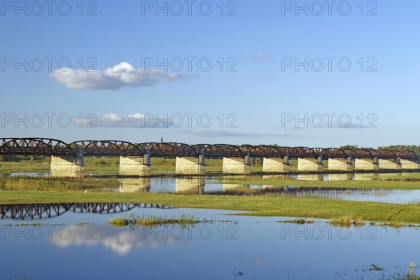 Long bridge over a calm river with reflections and green banks under a clear blue sky, autumn,