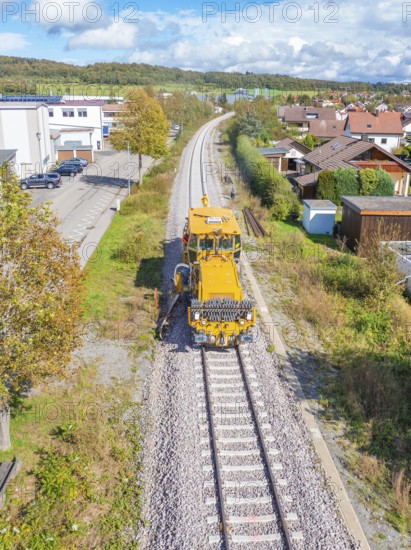 Yellow vehicle on ballast track in residential area under blue sky with clouds, tamping machine,