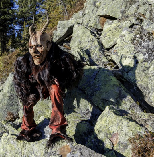 Person in devilish costume with mask and horns on rocks in front of a forest backdrop, Neukirchen