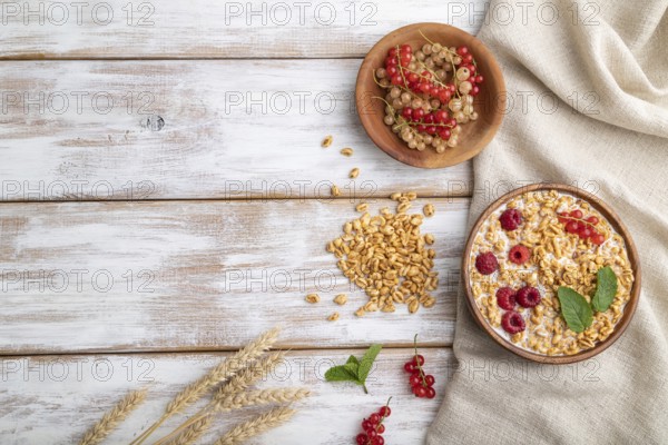 Wheat flakes porridge with milk, raspberry and currant in wooden bowl on white wooden background