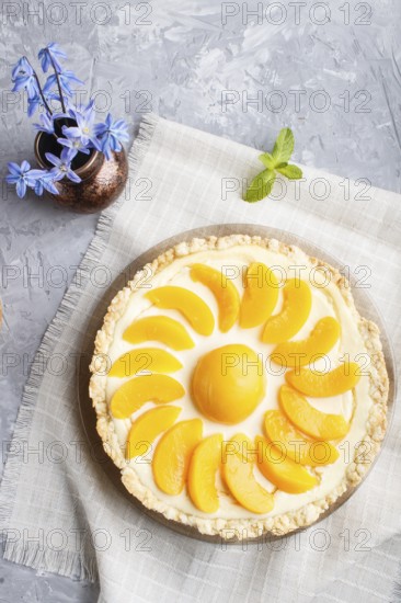 Round peach cheesecake and ceramic vase with blue flowers on a linen napkin on a gray concrete