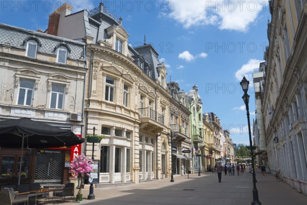 A lively city street with historic buildings under a blue sky on a sunny day, pedestrianised