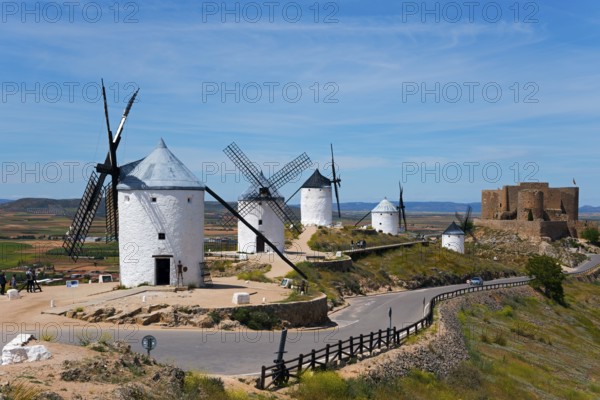 Several white windmills on a hill, with a castle in the background under a blue sky, Consuegra,