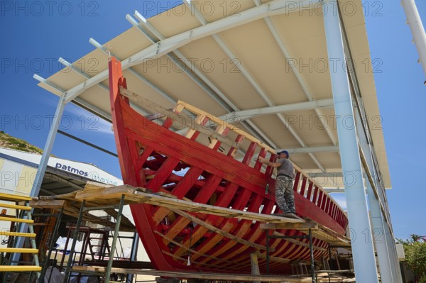 A partially built red wooden ship in a covered shipyard with a worker and blue sky, Patmos Marine,