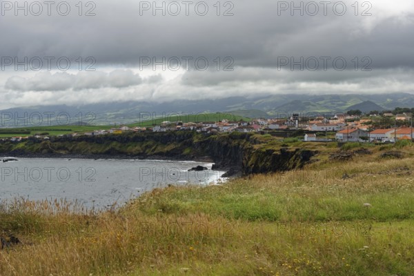 A village on the coast with green cliffs and a cloudy sky, Fenais da Luz, Sao Miguel Island,