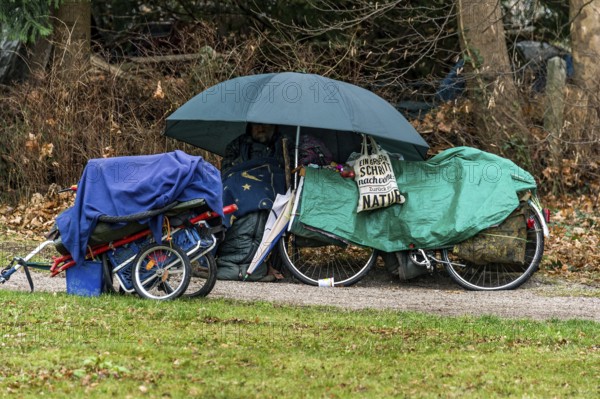 Homeless man in camp on park bench under umbrella with blanket and sleeping bag, bicycle covered