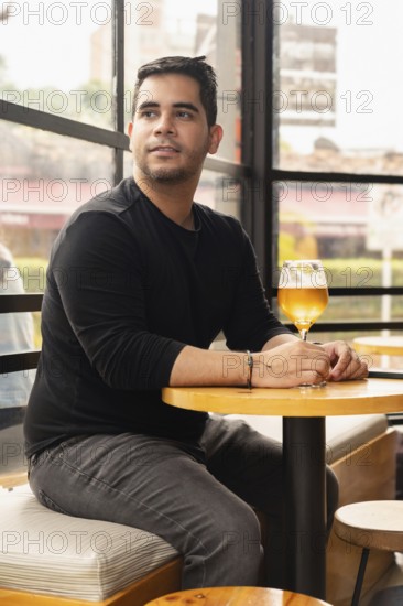 Handsome young Latino man sitting in a cafe with a glass of beer, gazing out the window. The cozy
