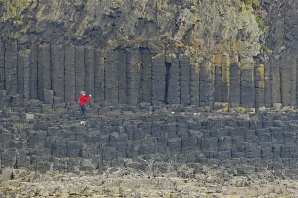 Person with red jacket standing in front of impressive basalt columns on a rocky cliff, Staffa,