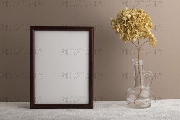 Brown wooden frame mockup with dried hydrangea in glass on beige paper background. Blank, vertical