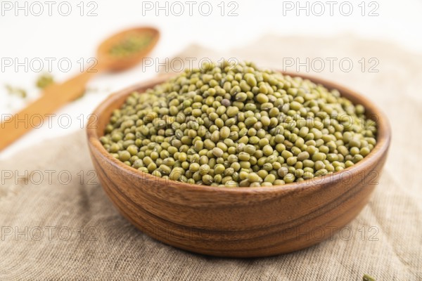 Wooden bowl with raw green mung bean and wooden spoon on a white background and linen textile. Side