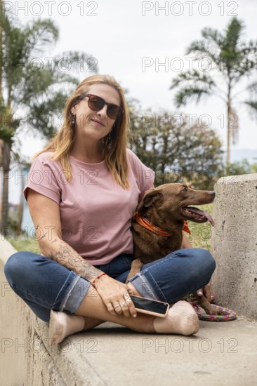 Middle-aged woman with sunglasses, sitting on a cement bench next to her dog, with palm trees in