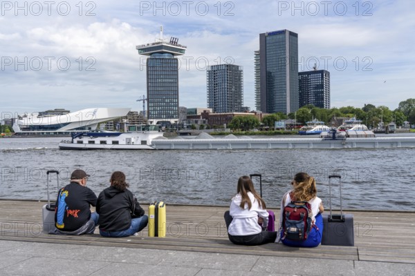 Tourists sitting at the back of Amsterdam Centraal railway station, on the promenade of the river